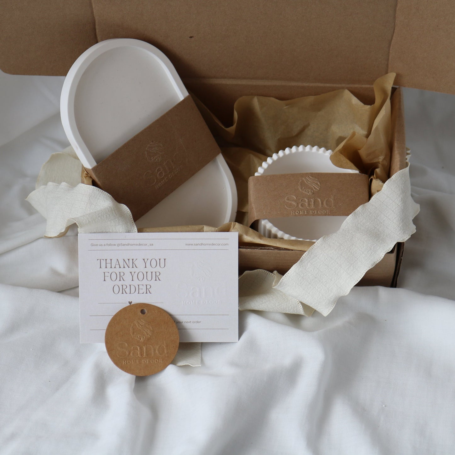 Gift box from Sand Home Decor featuring a white oval dish and a trinket tray, both wrapped in kraft paper bands. Accompanied by a 'Thank You for Your Order' card and a circular kraft paper tag with the Sand Home Decor logo, set on a soft white fabric background.