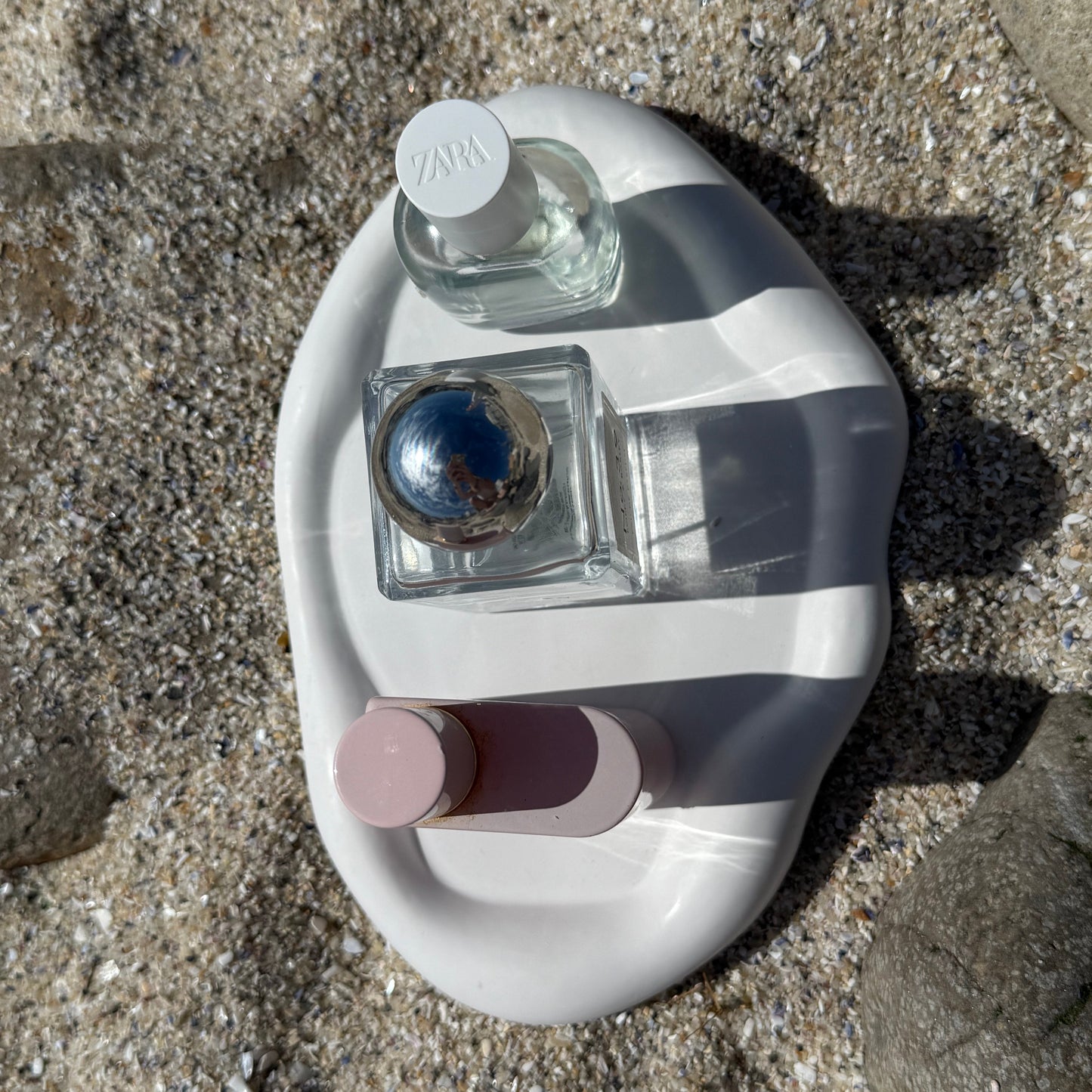Birds eye view of an organic-shaped handmade white tray made from eco resin, styled on beach sand with natural stones and perfume bottles.