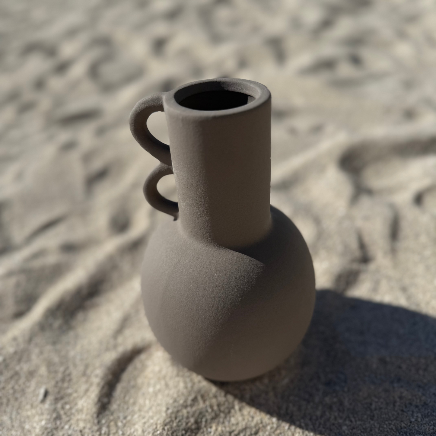 Matte mocha-colored ceramic vase with a spherical base and two sculptural loop handles on a cylindrical neck sitting on a sandy beach.