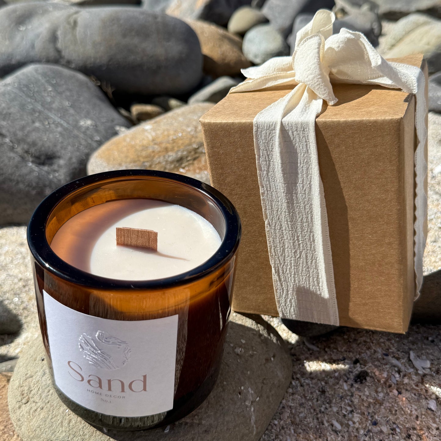 Large scented soy candle in an amber glass jar with a beach setting as the background and packaging displayed next to it.