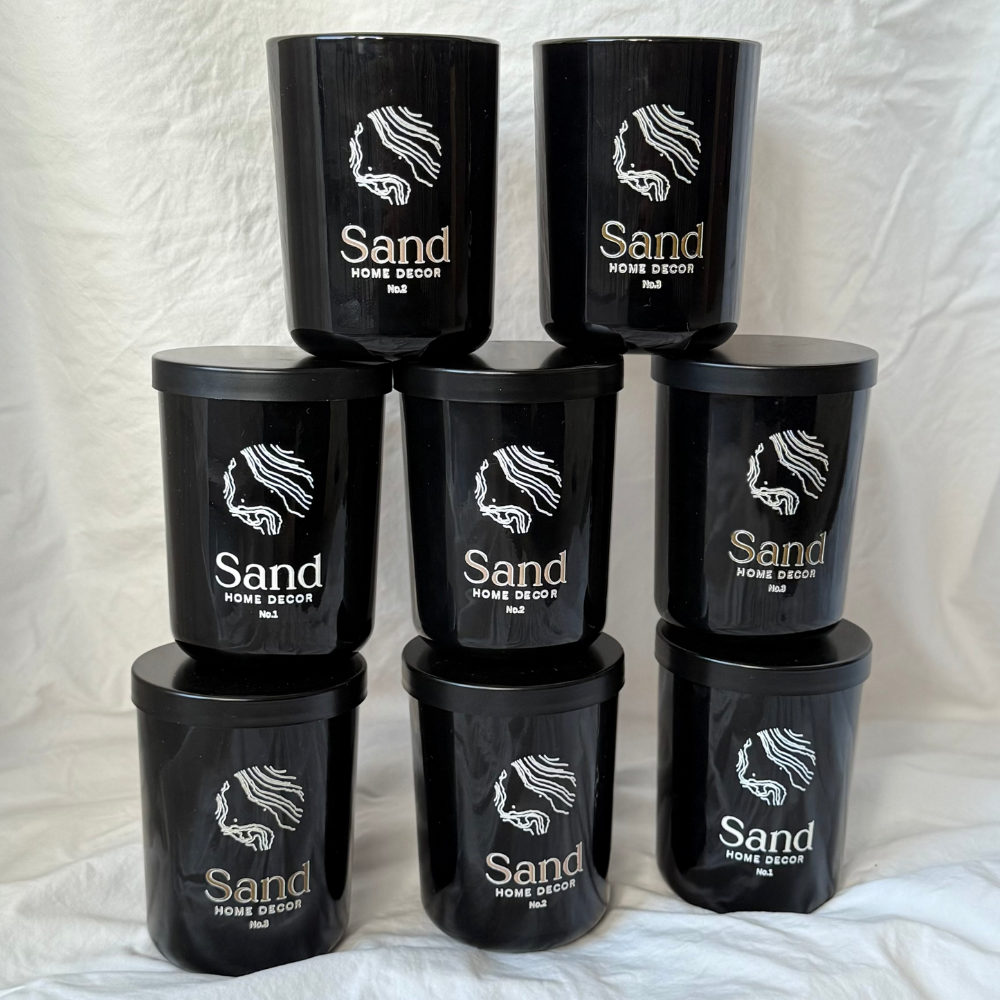 Black candle containers with 'Sand Home Decor' branding on a white fabric background