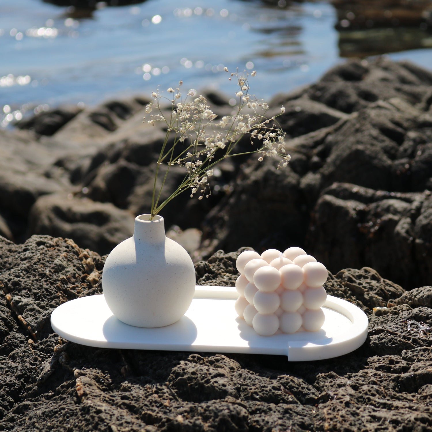 A decorative bubble cube candle in off white, displayed outdoors on a white tray alongside a minimalist ceramic vase with delicate white flowers. The scene features a natural rocky backdrop and a blurred ocean view.