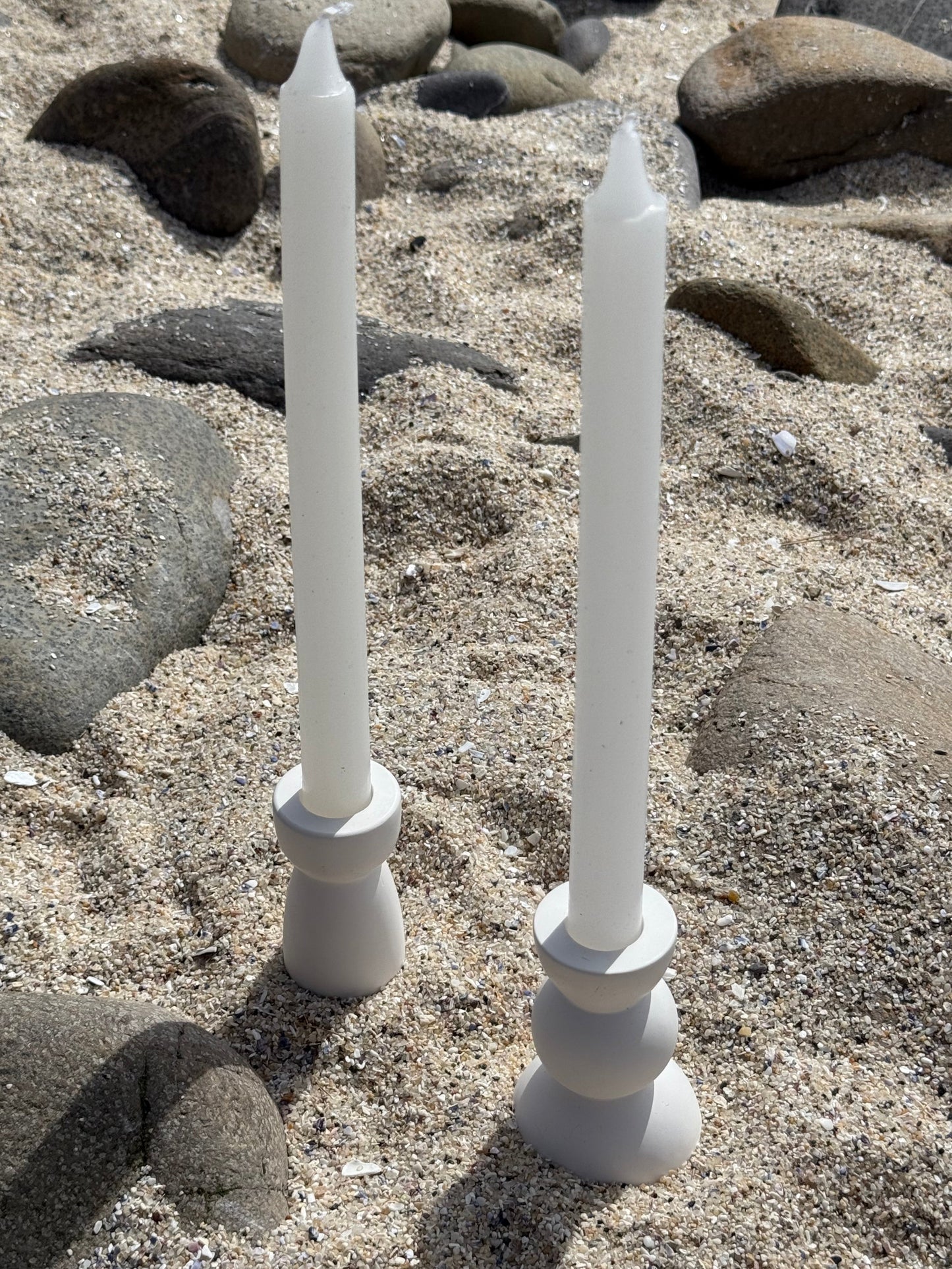 Two modern white candle holders  made from eco resin with candles, placed on a sandy beach with natural stones in the background.