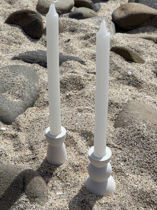 Two modern white candle holders  made from eco resin with candles, placed on a sandy beach with natural stones in the background.