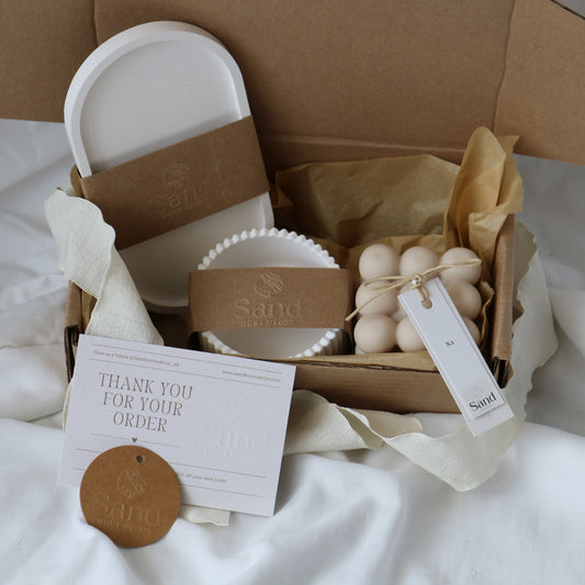 Gift box from Sand Home Decor featuring a white trinket tray, ribbed trinket dish and a decorative candle, wrapped in branded kraft paper bands. Accompanied by a 'Thank You for Your Order' card and a circular kraft paper tag with the Sand Home Decor logo, set on a soft white fabric background.