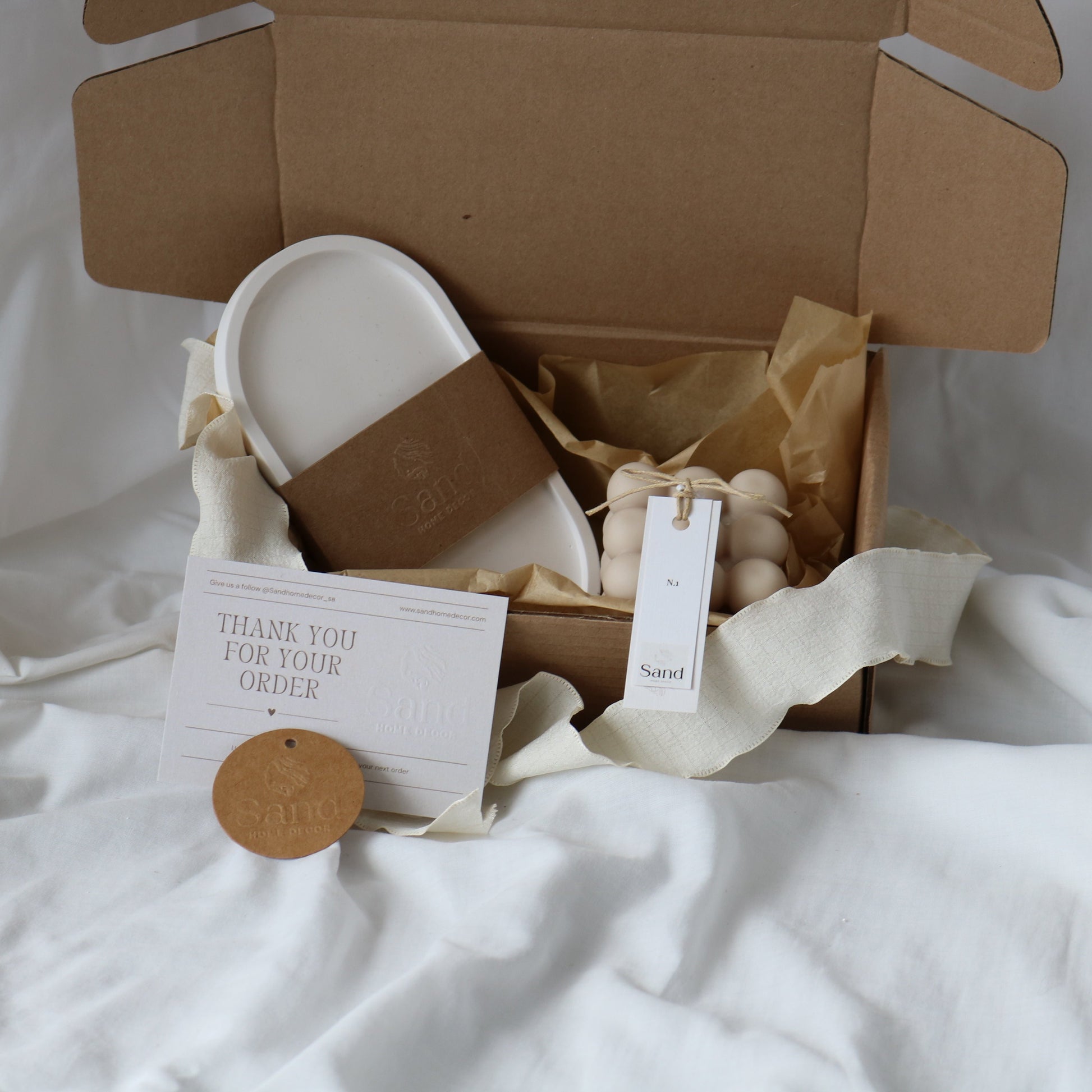 Gift box from Sand Home Decor featuring a white handmade trinket tray and a decorative bubble candle, both wrapped in branded kraft paper bands. Accompanied by a 'Thank You for Your Order' card and a circular kraft paper tag with the Sand Home Decor logo, set on a soft white fabric background.