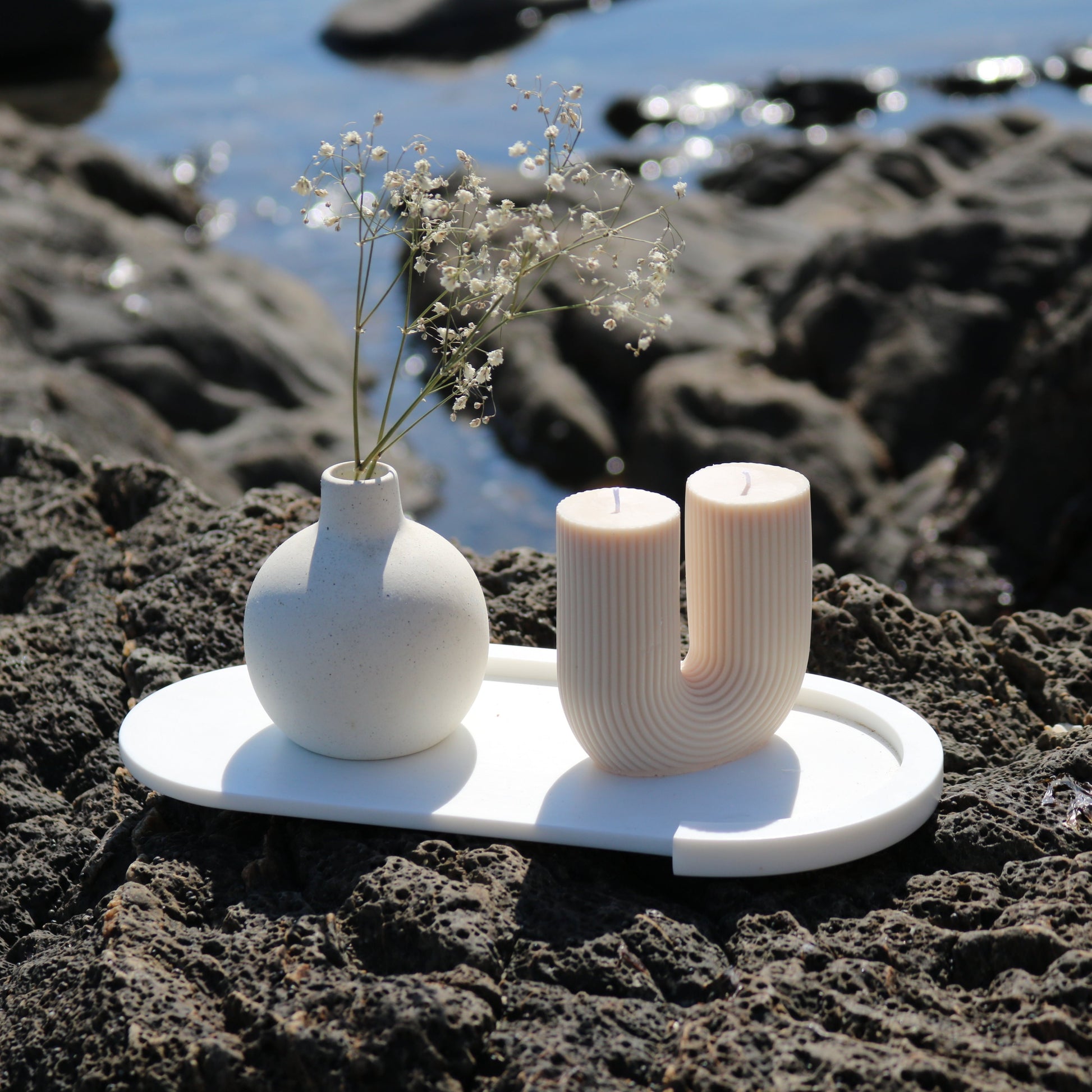 A decorative U Shaped candle in soft white, displayed outdoors on a white tray alongside a minimalist ceramic vase with delicate white flowers. The scene features a natural rocky backdrop and a blurred ocean view.