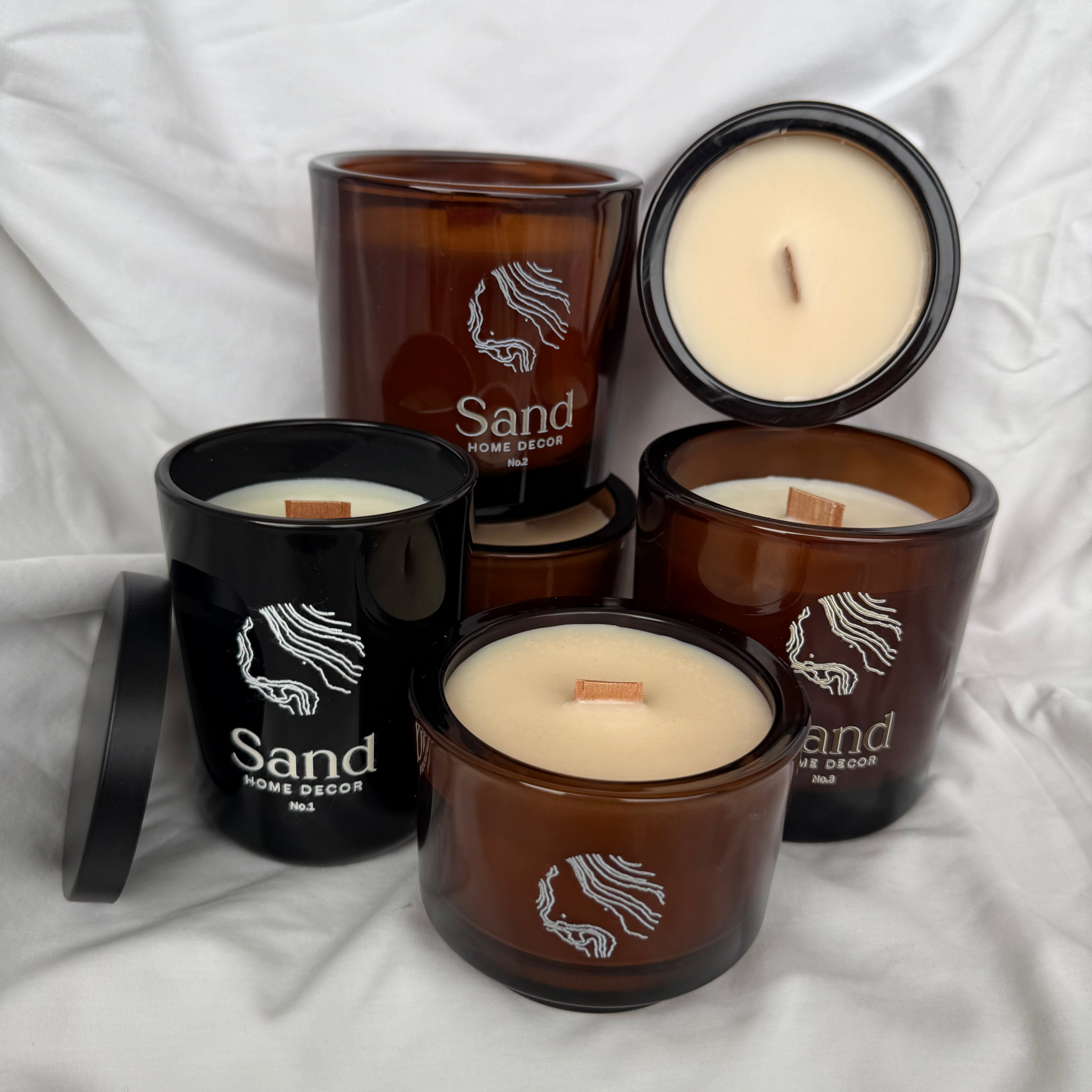A group of Sand Home Scents soy candles in amber and black glass jars, with one jar shown open to reveal the cream-colored wax and wick, arranged on a white fabric background.