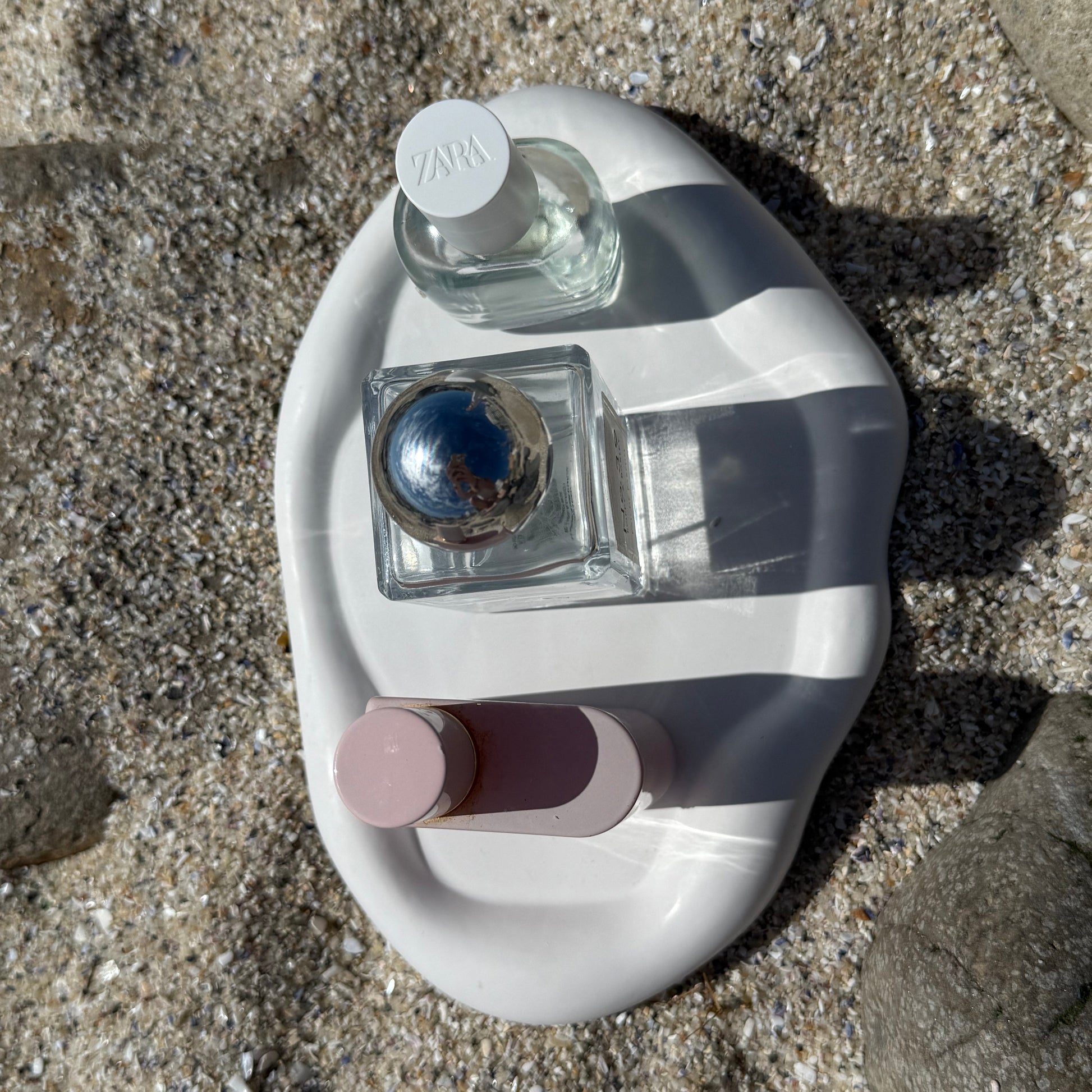 Birds eye view of an organic-shaped handmade white tray made from eco resin, styled on beach sand with natural stones and perfume bottles. 

