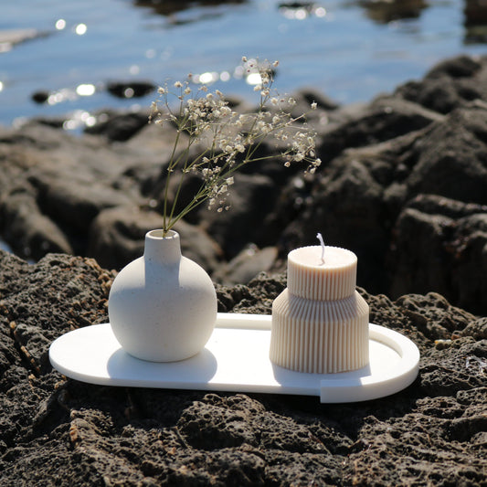 A decorative tiered handmade candle in off white, displayed outdoors on a white tray alongside a minimalist ceramic vase with delicate white flowers. The scene features a natural rocky backdrop and a blurred ocean view.