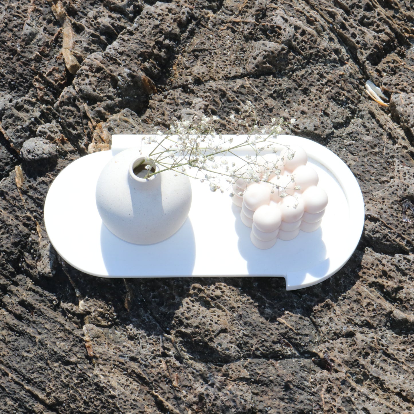 A decorative bubble cube candle in soft white, displayed outdoors on a white tray alongside a minimalist ceramic vase with delicate white flowers. The scene features a natural rocky background,