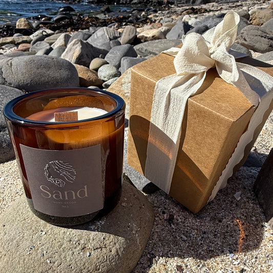 Large scented soy candle in an amber glass jar with a beach setting as the background and packaging displayed next to it.