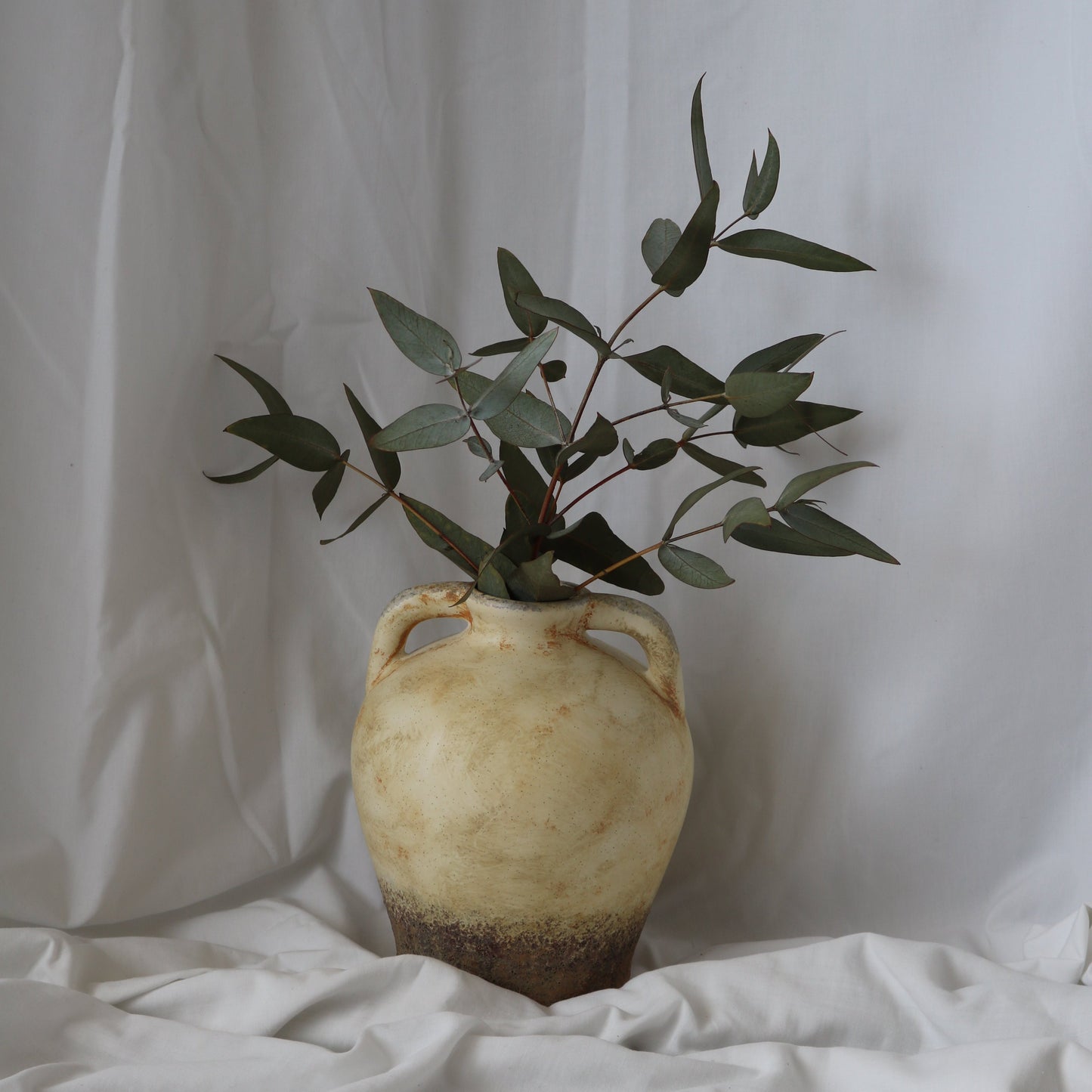 Rustic Mediterranean round vase with a dark brown bottom and rustic beige top half. With a white linen back drop. 