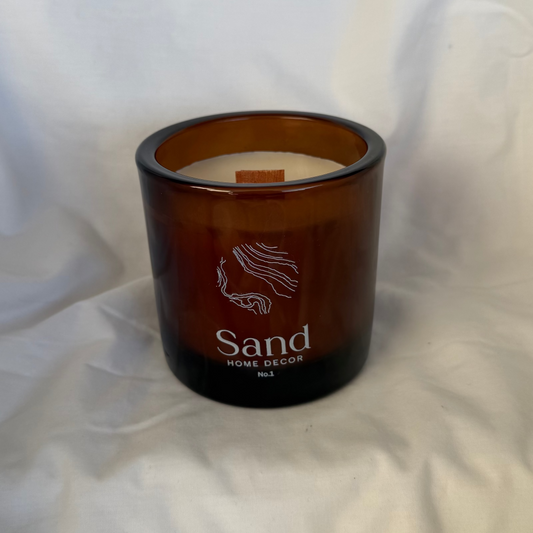 An amber glass jar candle with white wax and a wooden wick, featuring the brand name 'Sand' and scent 'Home Sweet Home' printed on the front, set against a draped white fabric background.