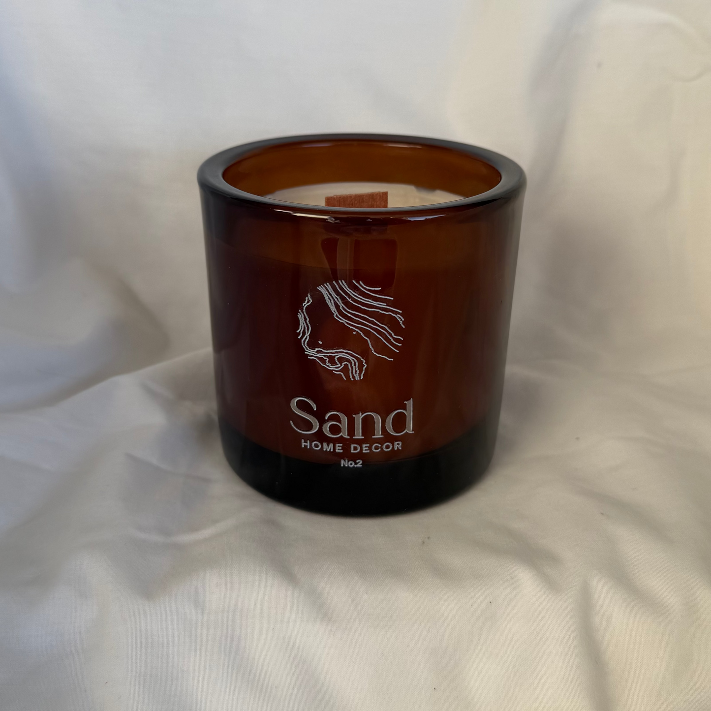 An amber glass jar candle with white wax and a white printed logo and text reading 'Sand Home Decor' on a draped white fabric background.