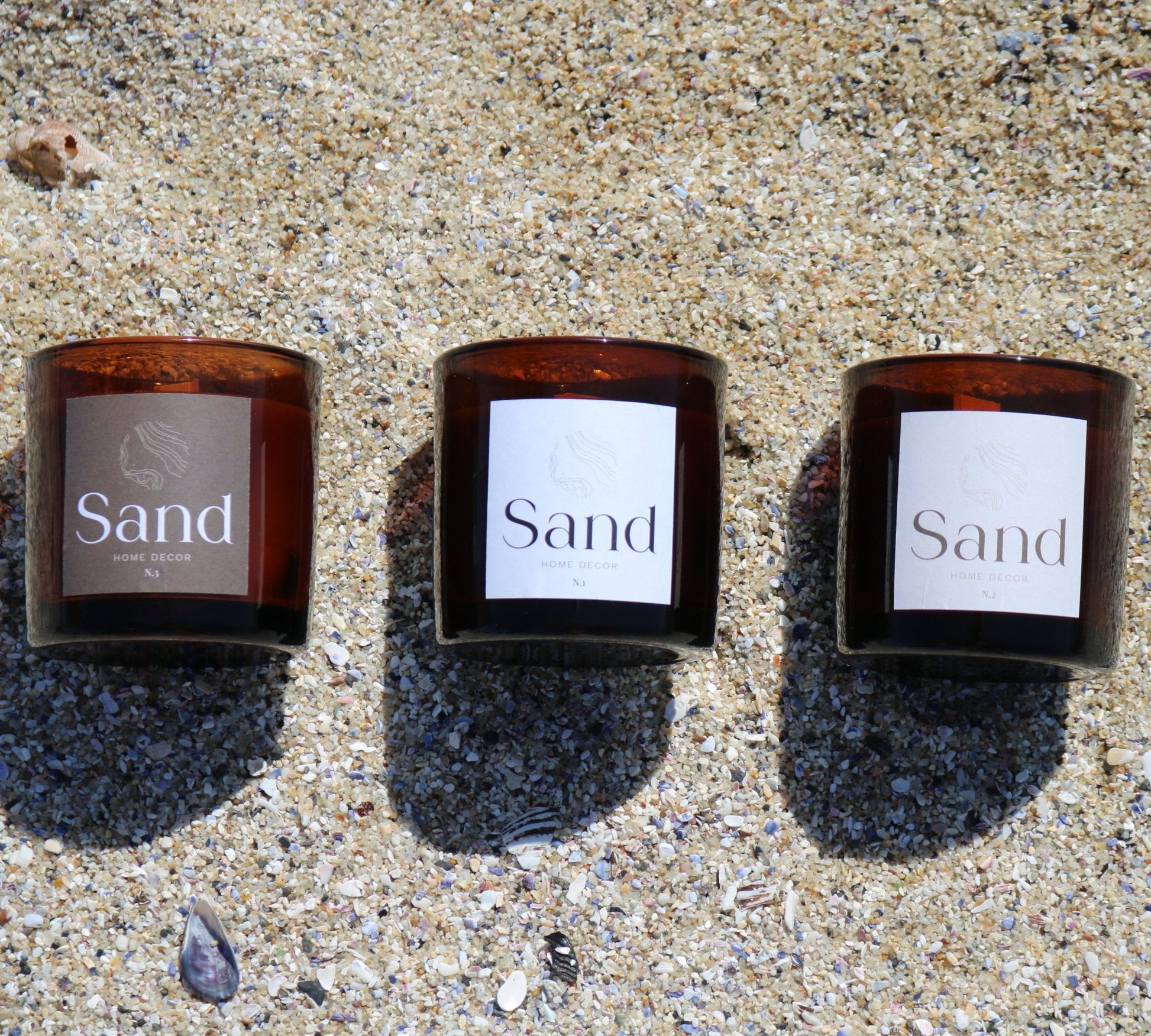 Three large soy scented luxury handmade candles in an amber glass jar lined up on beach sand. 