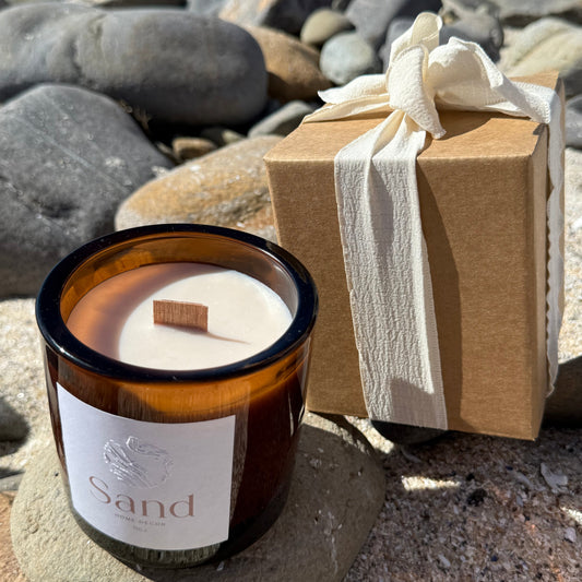 Large scented soy candle in an amber glass jar with a beach setting as the background and packaging displayed next to it. 
