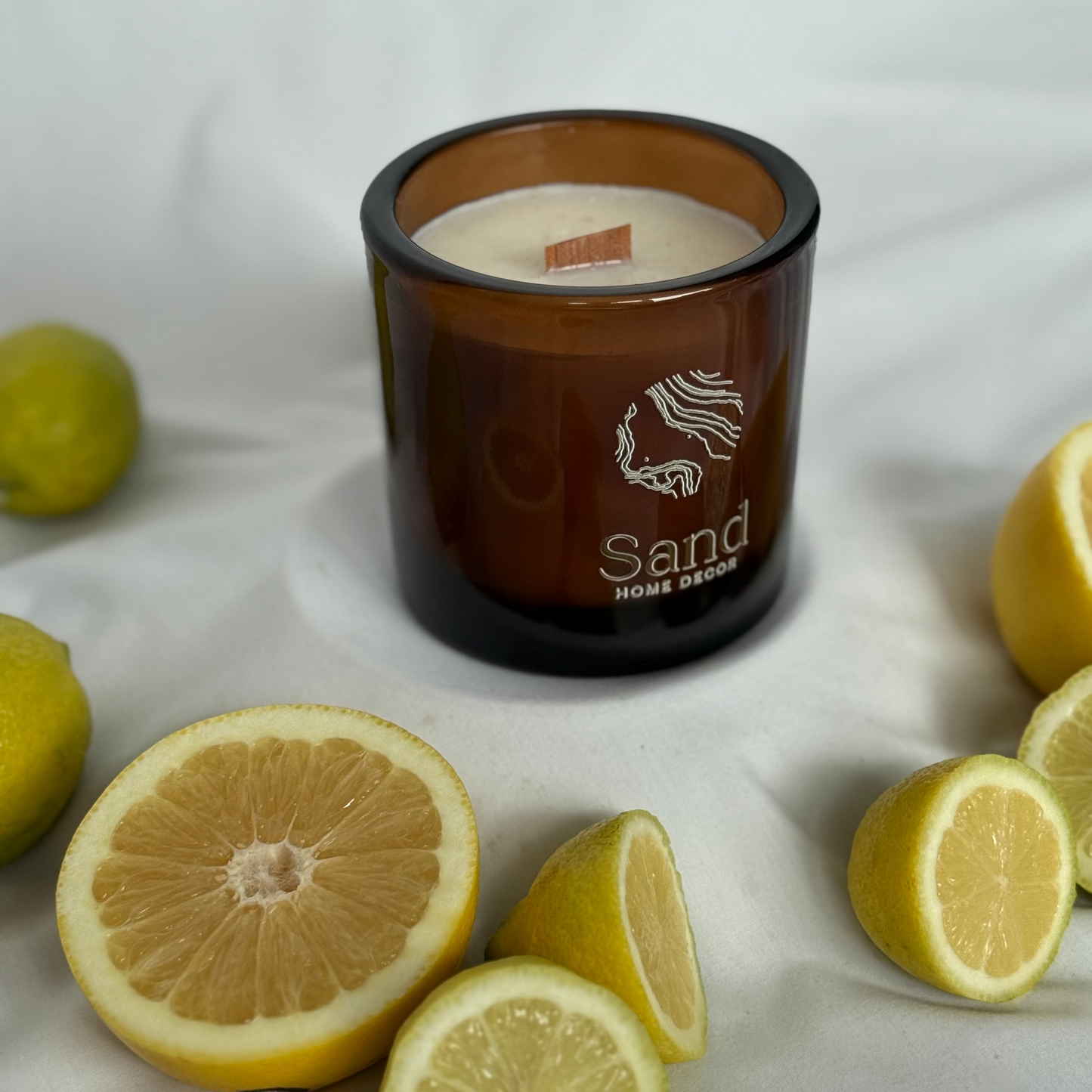 Candle with 'Sand Home Decor' branding surrounded by lemons on a white fabric background