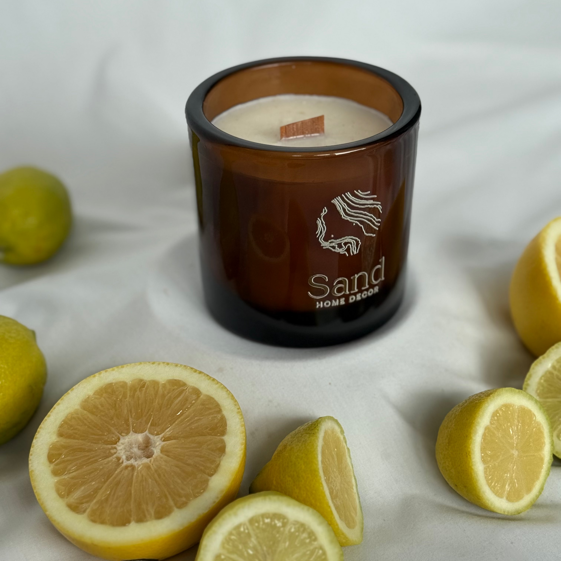Candle with 'Sand Home Decor' branding surrounded by lemons on a white fabric background