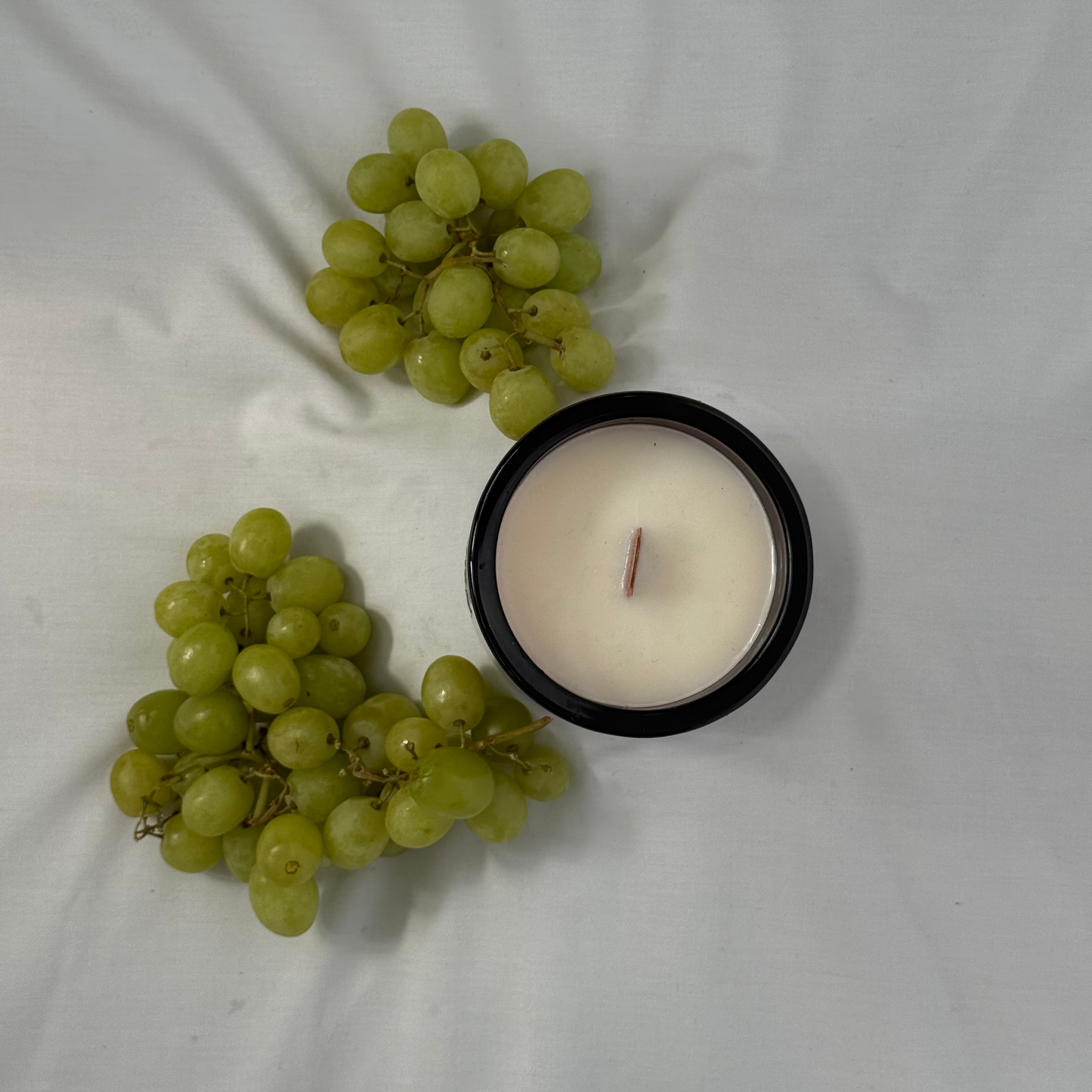 Two bunches of green grapes and a white candle in a black holder on a white fabric background