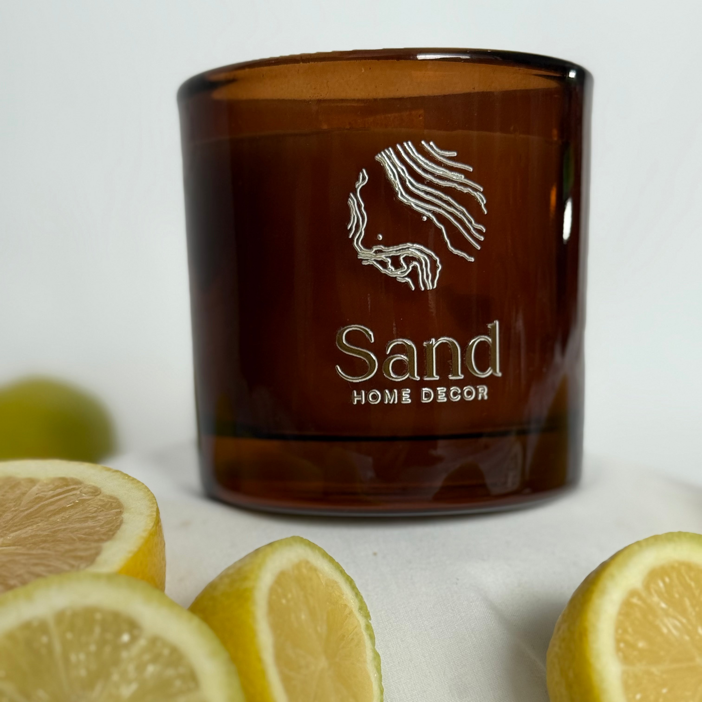 Brown candle with 'Sand Home Decor' branding surrounded by sliced lemons on a light background