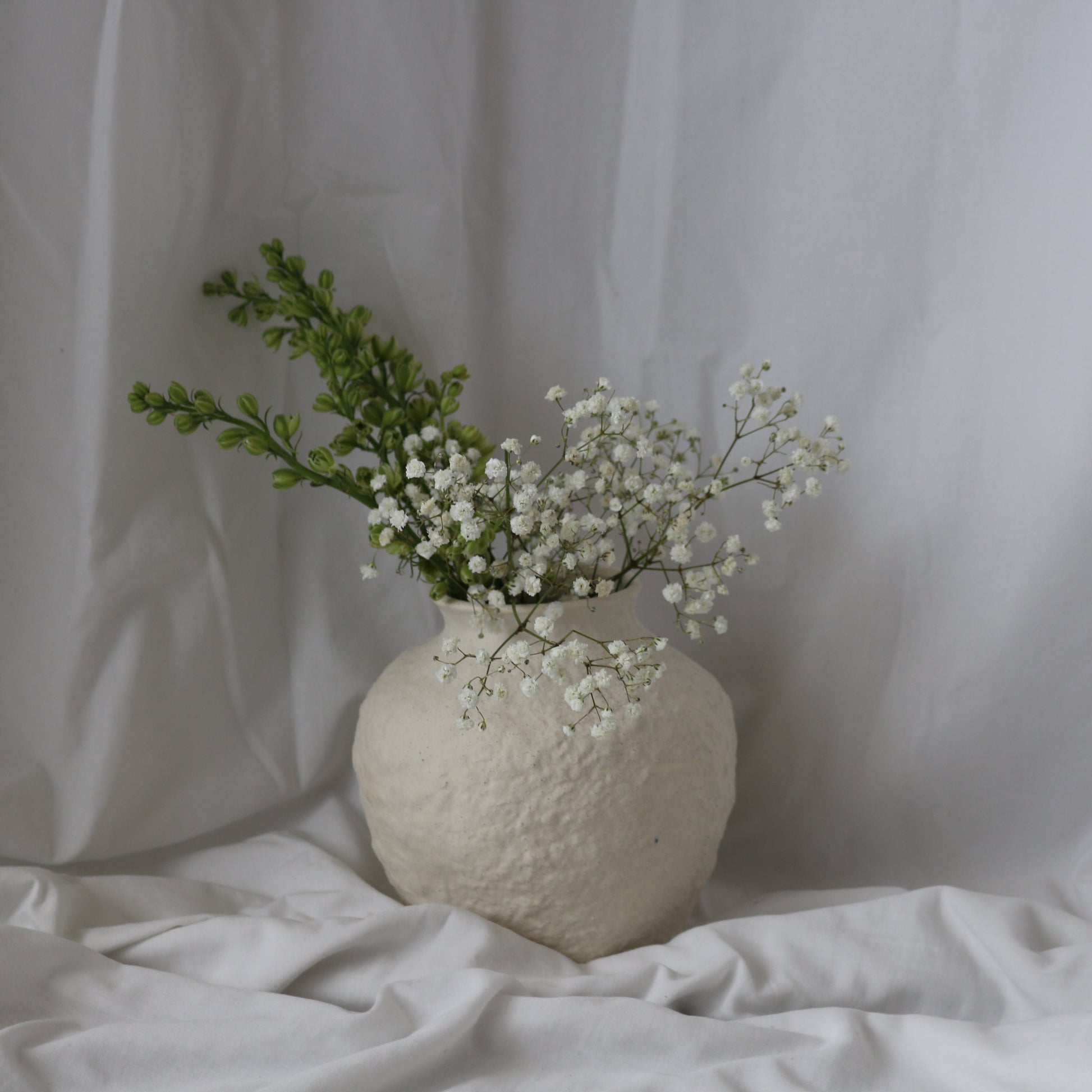 Very textured white ceramic round vase with foliage with a white linen background.